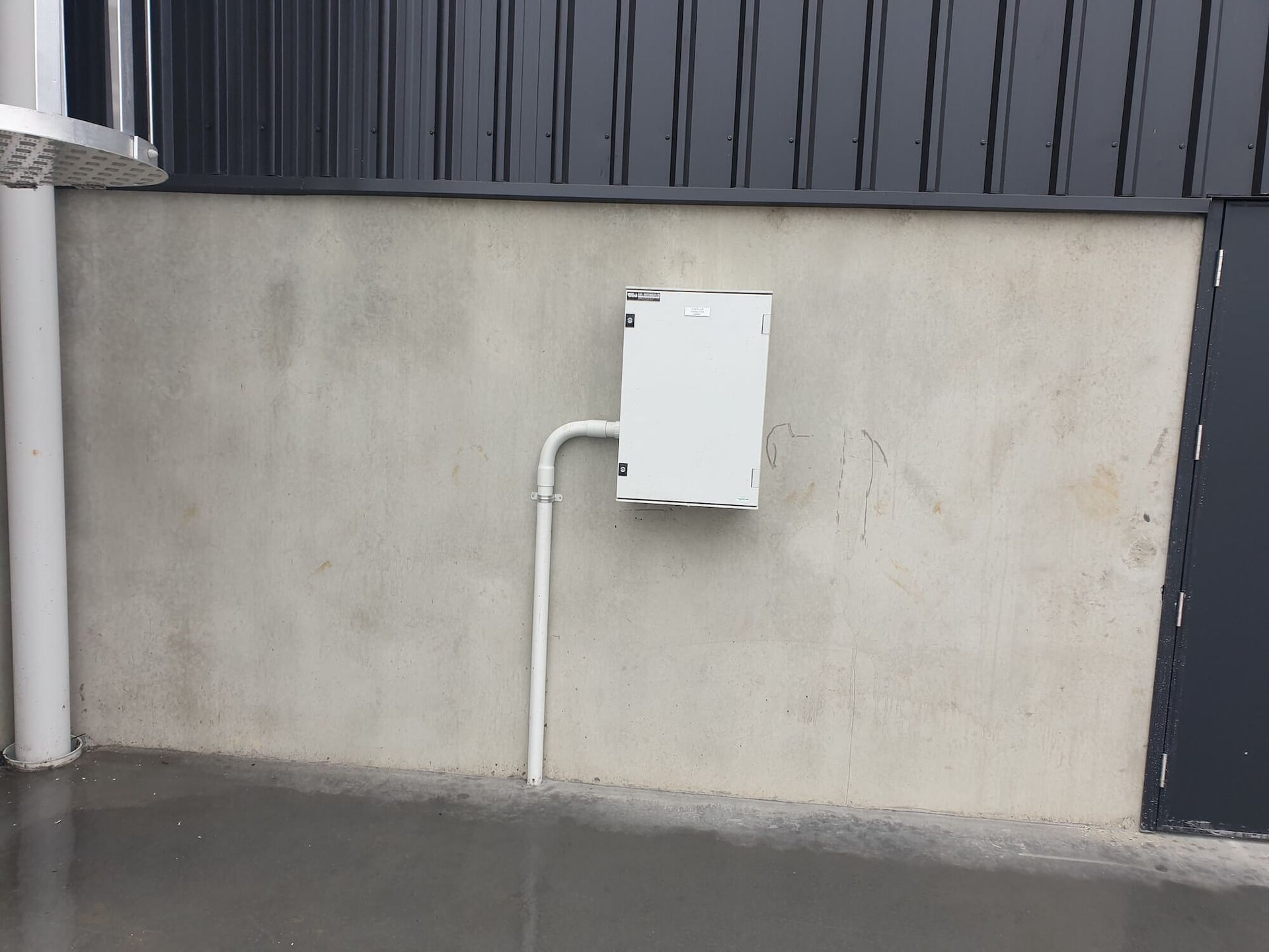 White electrical box on a gray concrete wall next to a dark metal building and a white pipe.