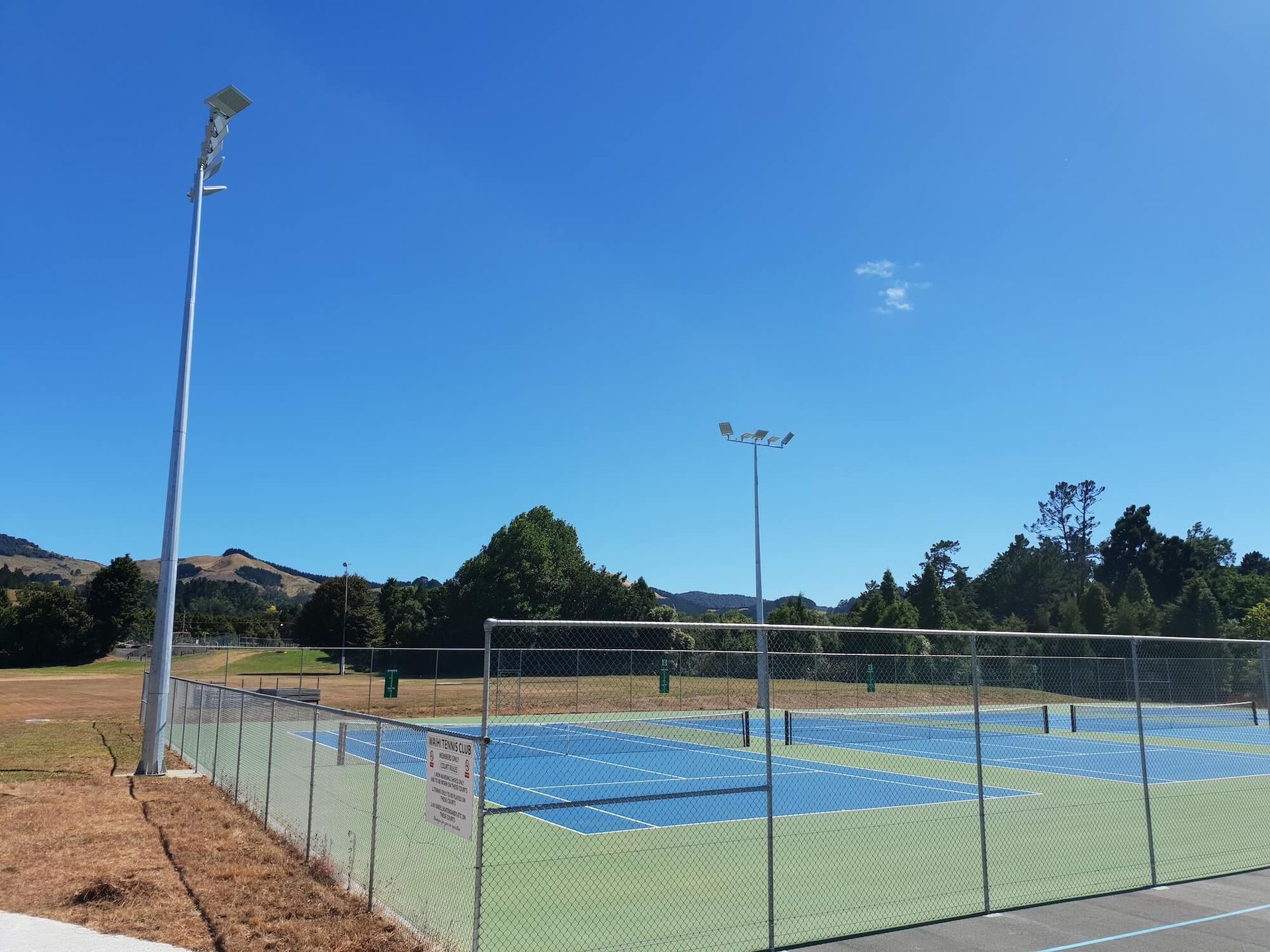 Tennis courts behind a chain-link fence under a clear, bright blue sky. Tall light poles are near the courts, with green and blue court surfaces.