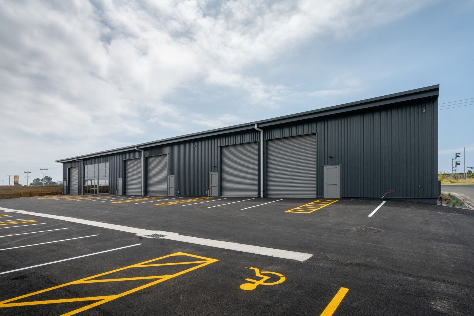 Dark gray industrial building with roll-up doors, asphalt parking lot with yellow markings, including a handicapped parking space.