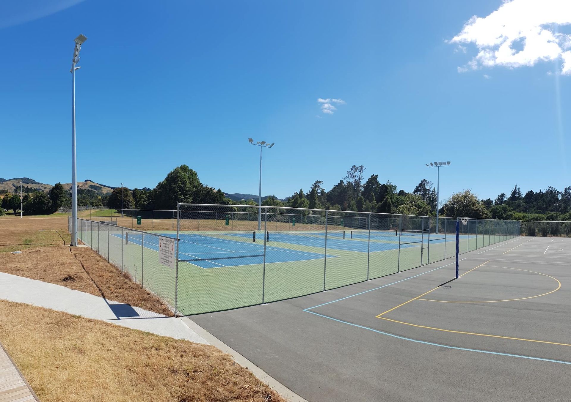 Outdoor sports court with blue and green surface, surrounded by a fence and pathway, under a blue sk