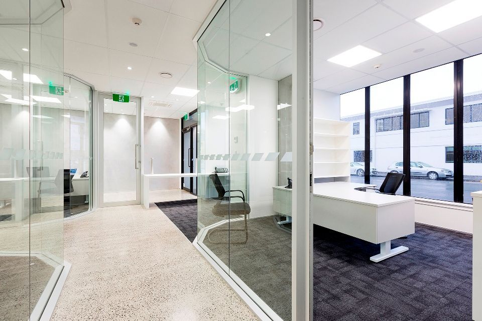 Modern office interior with glass walls, a desk, and a hallway. White and gray color scheme with natural light.