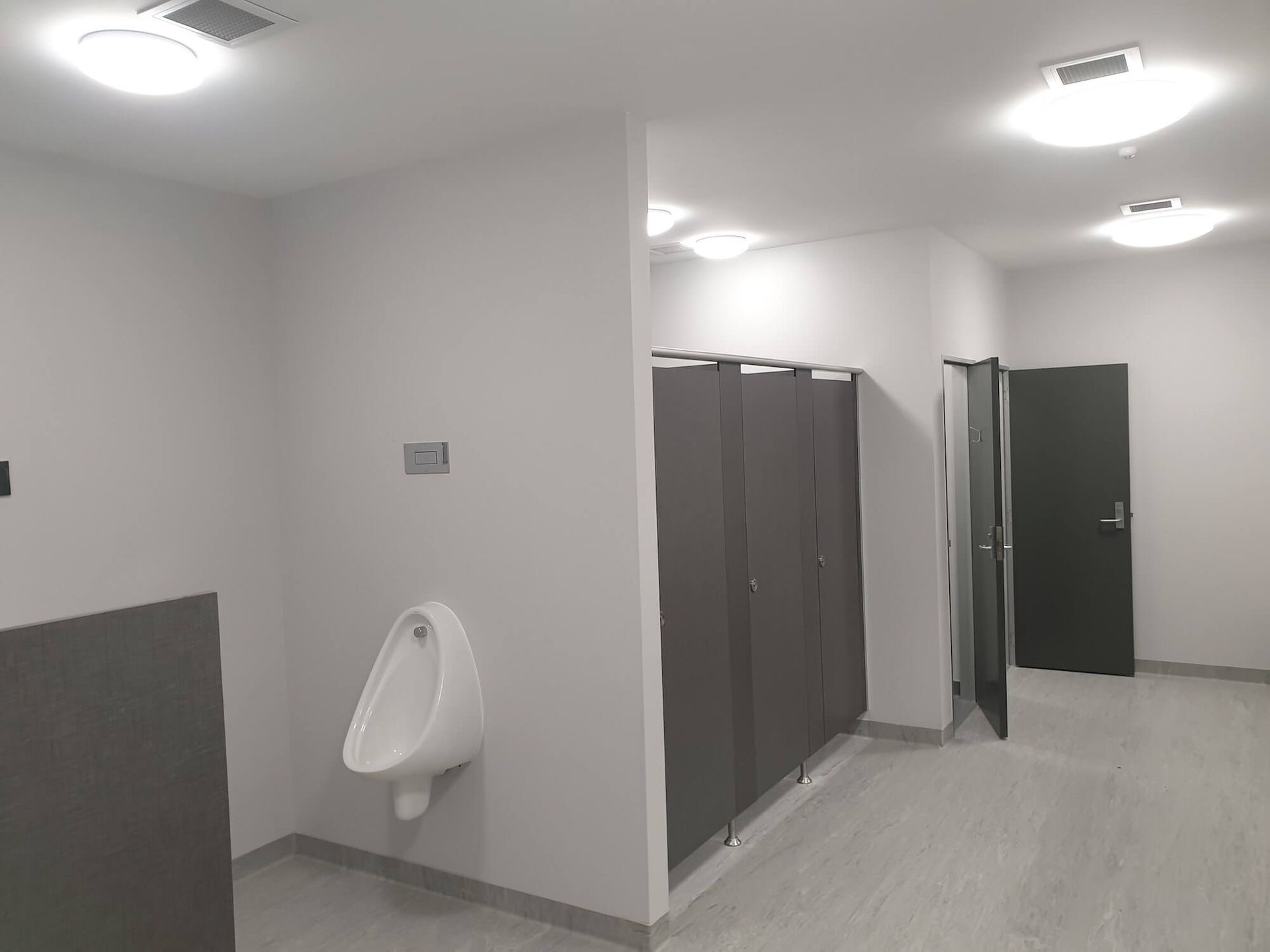 A public restroom featuring stalls and urinals. White walls, grey stall doors, and bright ceiling lights.