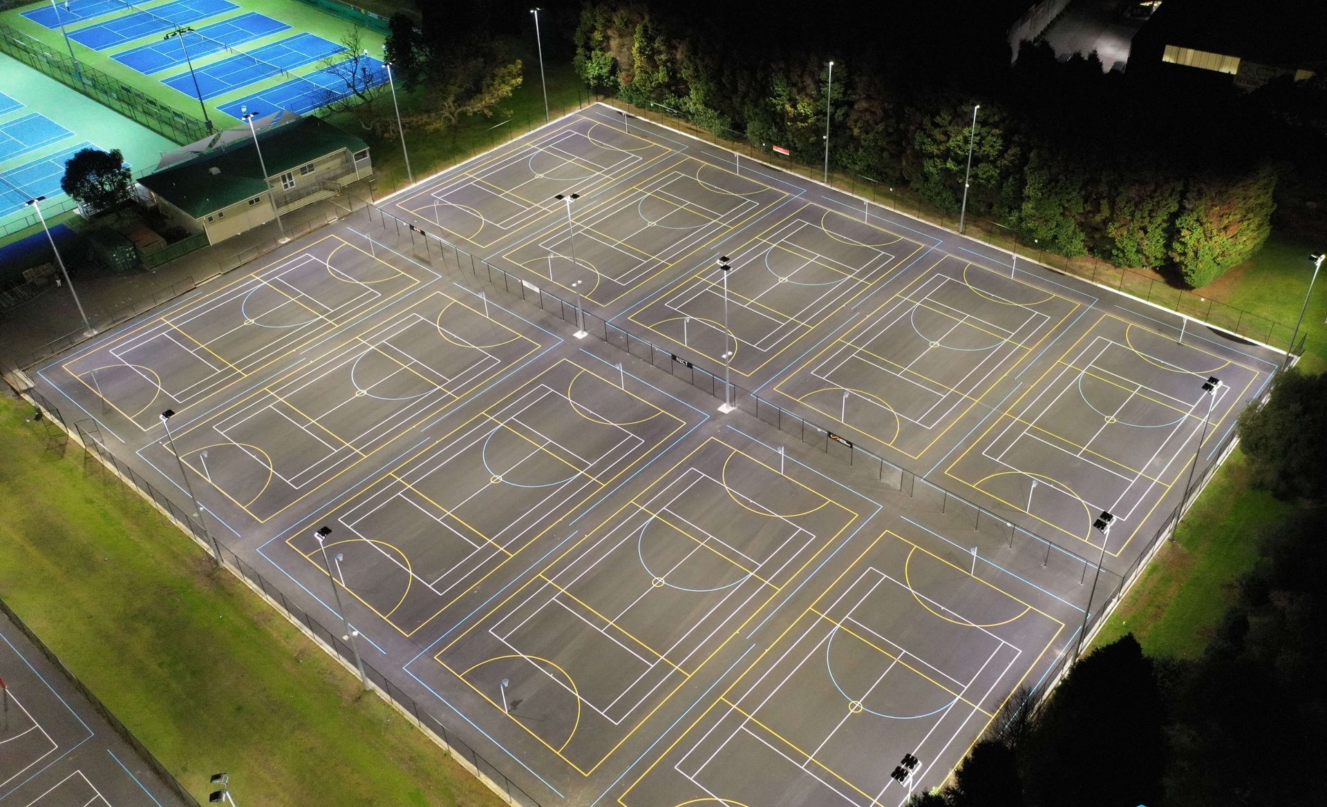 Aerial view of a brightly lit multi-sport court at night, with white lines marking various playing areas, surrounded by green grass.