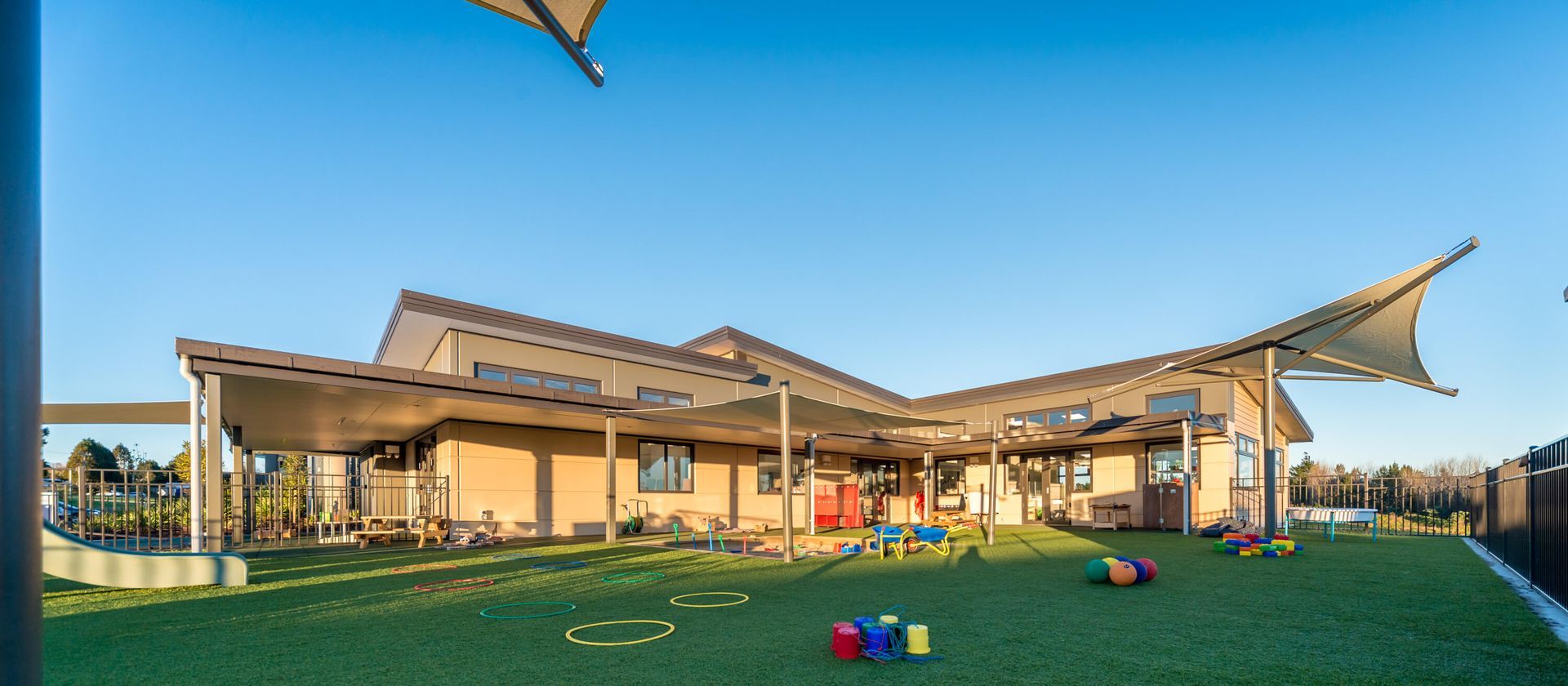 A daytime view of a daycare building with a covered outdoor play area and green artificial turf. Colorful toys are scattered on the grass.