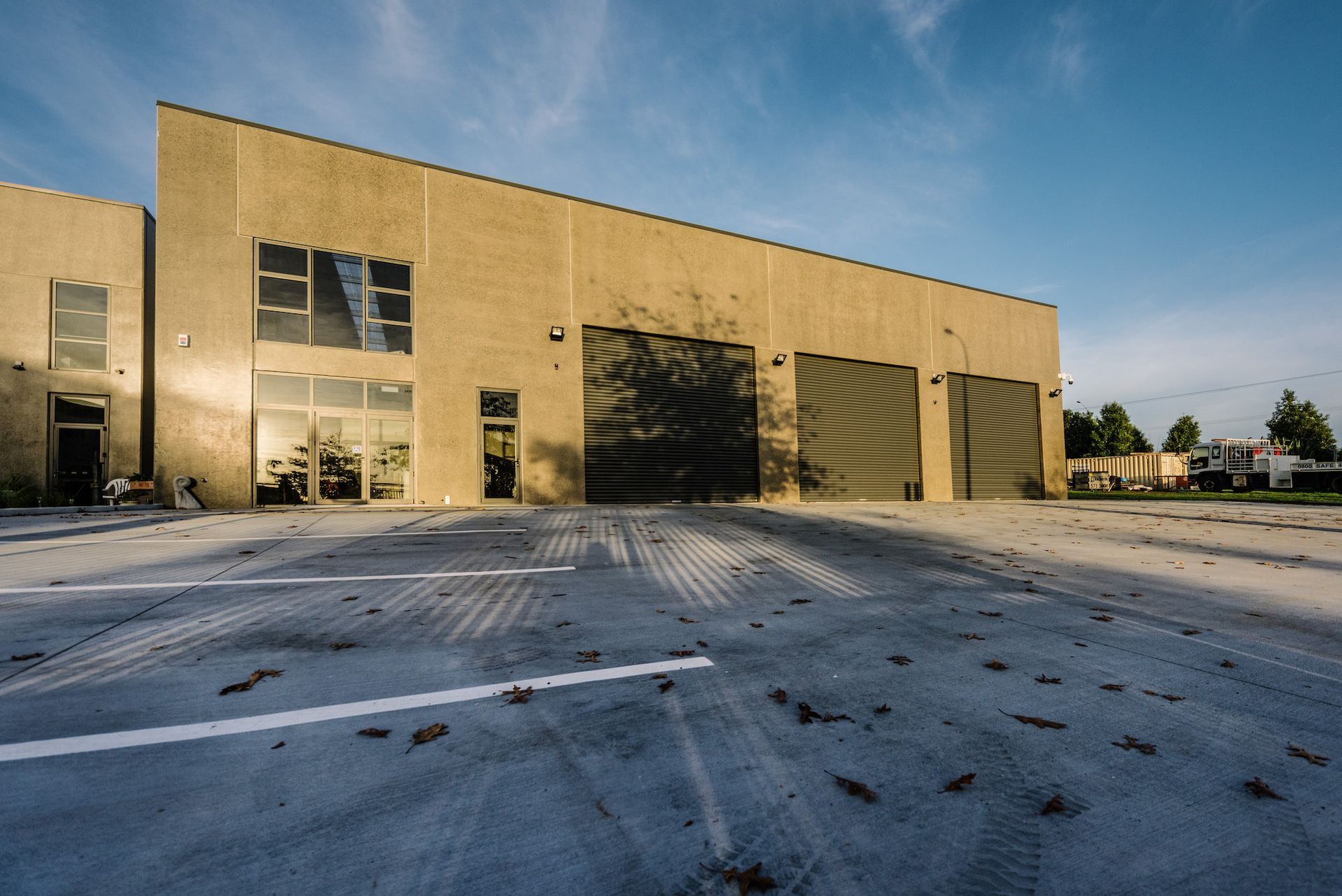 A tan industrial building with large garage doors and a paved lot under a blue sky, casting long shadows.