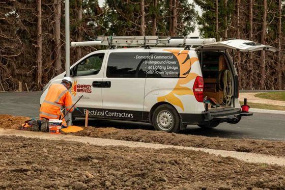 Worker in orange vest using a tool near a white work van with ladder on top and open back.
