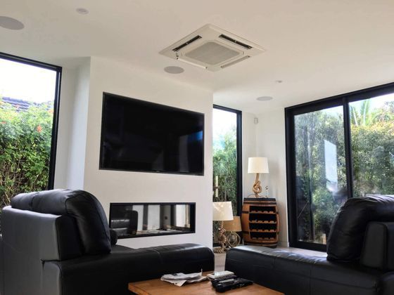 Modern living room with black leather sofas, large TV, fireplace, and windows overlooking greenery.