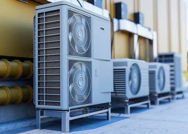 Several air conditioning units lined up against a beige wall with yellow pipes, a blue-gray ground,