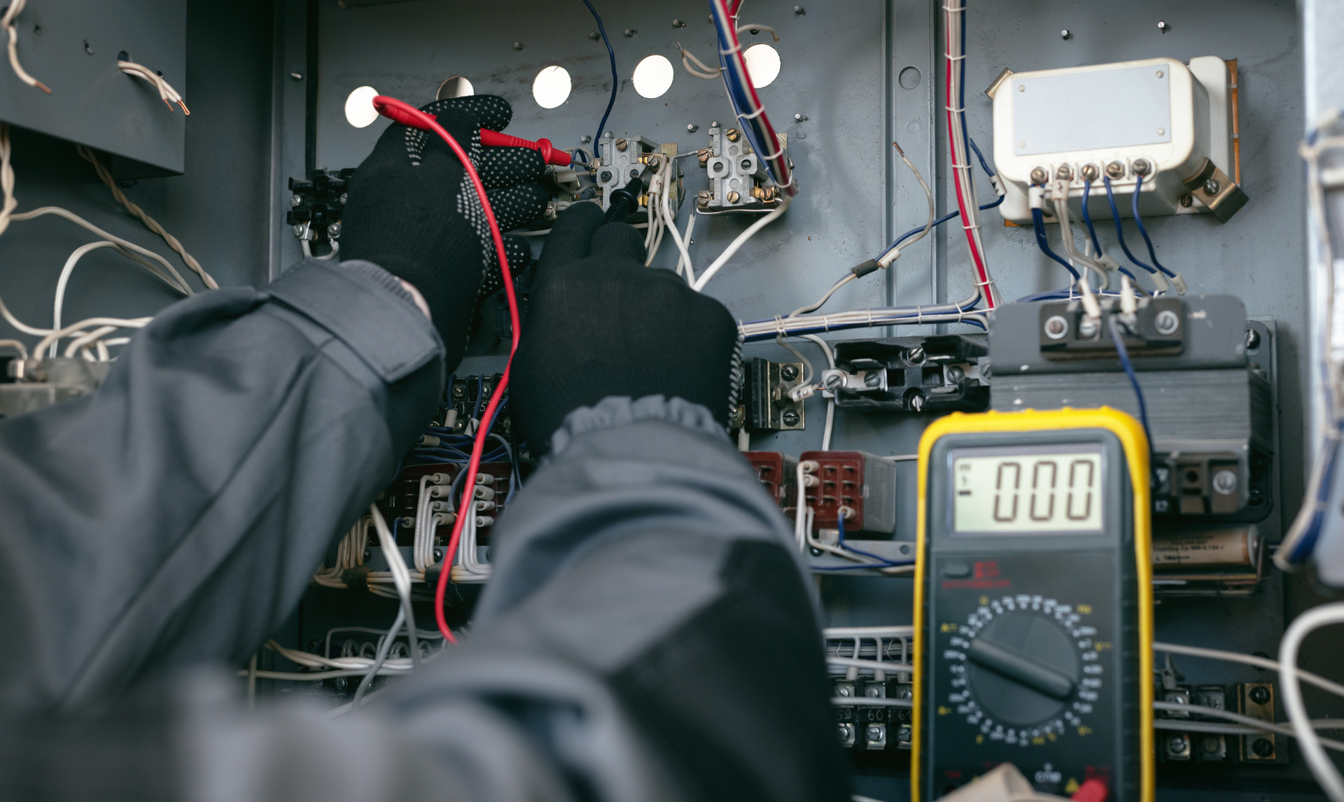 Electrician in black gloves using a multimeter to test wires in a control panel.