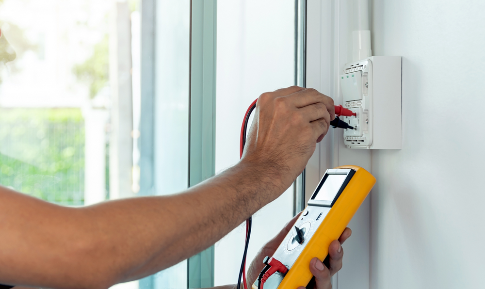 Electrician using a multimeter to test a wall outlet, indoors with natural light.