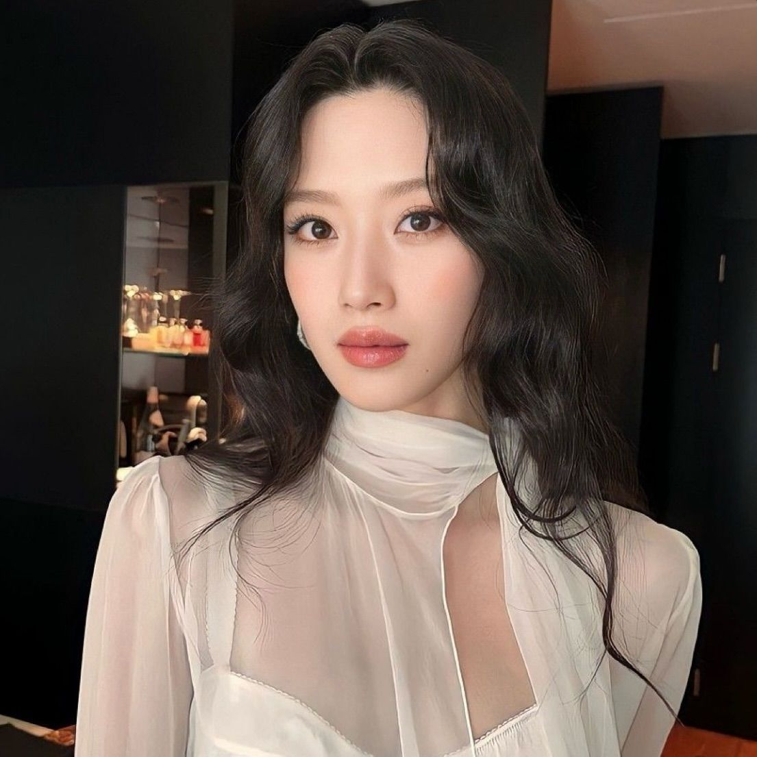 Woman with dark wavy hair, wearing a sheer white top. She's looking at the camera with a neutral expression.