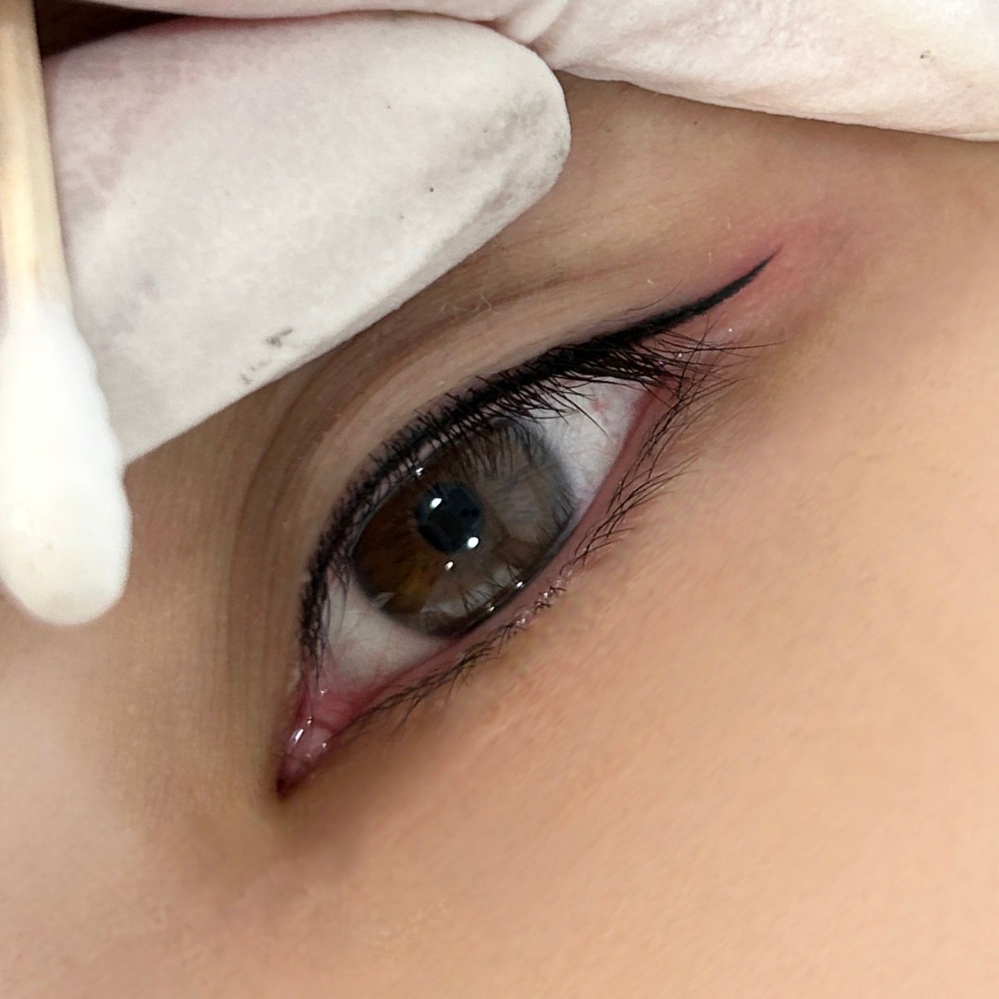 Close-up of an eye with freshly applied black eyeliner; a gloved hand holds a cotton swab.