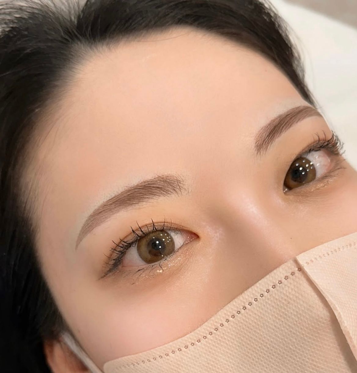 Close-up of a person's eyes, eyebrows, and face mask, showcasing microbladed brows and contact lenses.
