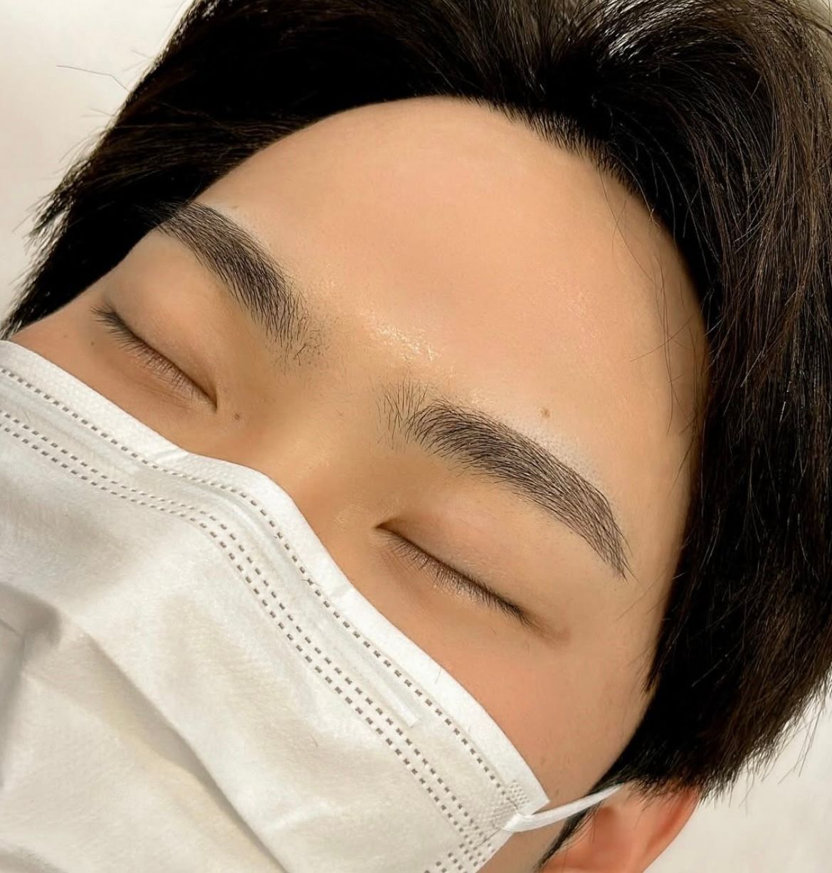 Close-up of a person wearing a mask, eyes closed, freshly microbladed eyebrows.