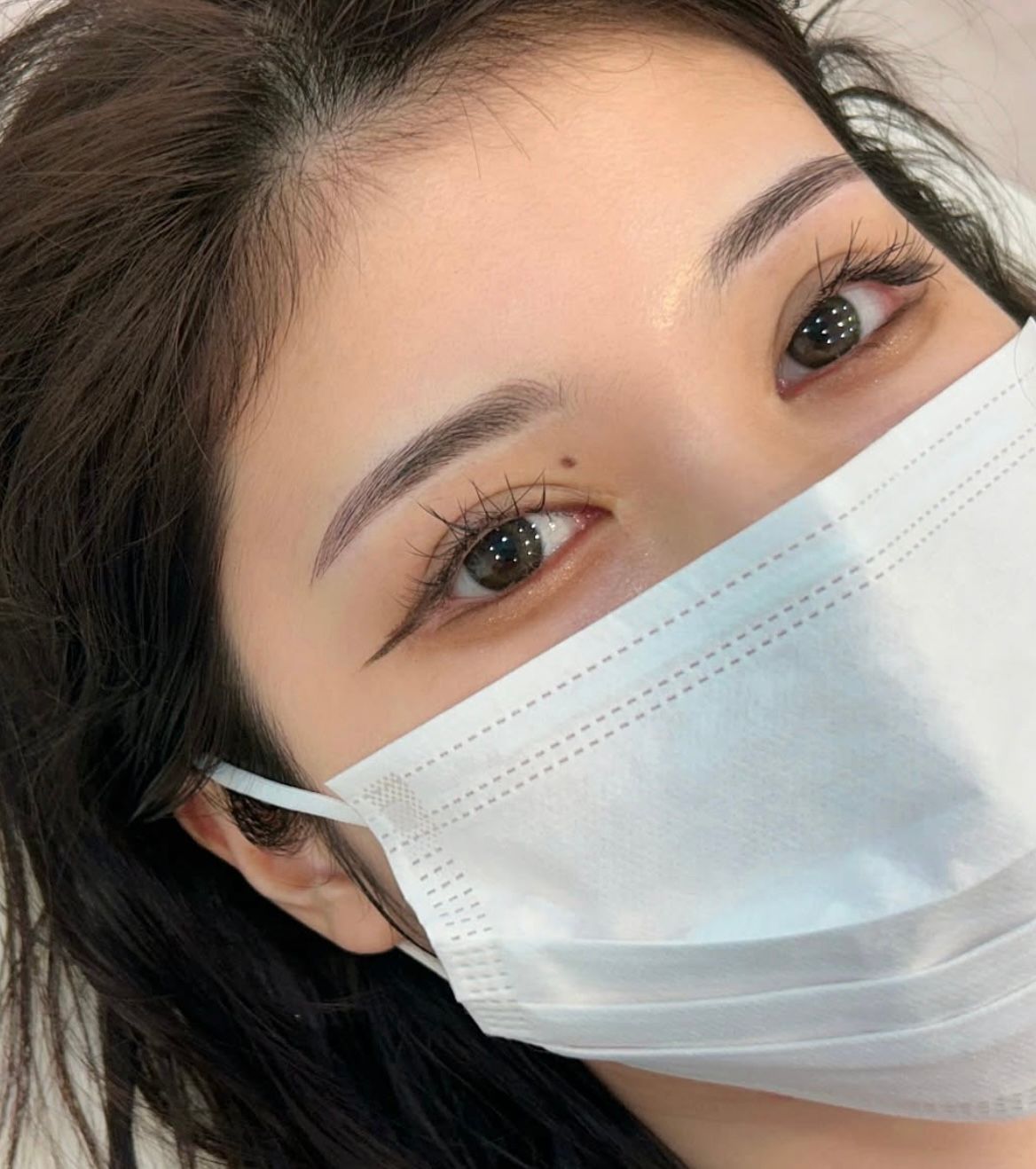 Woman with a face mask, eyeliner, and eyebrow tattoo, looking at the camera.