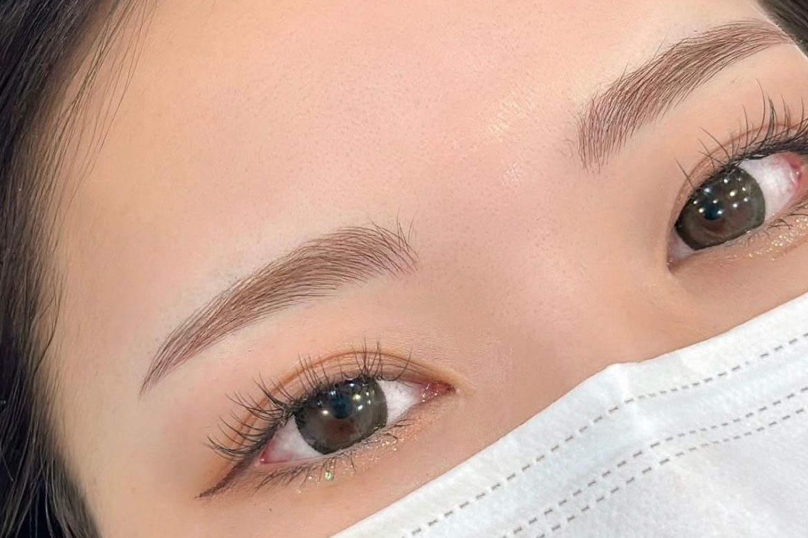 Close-up of eyes, face partially obscured by mask, showing filled-in eyebrows, long lashes, and light brown irises.