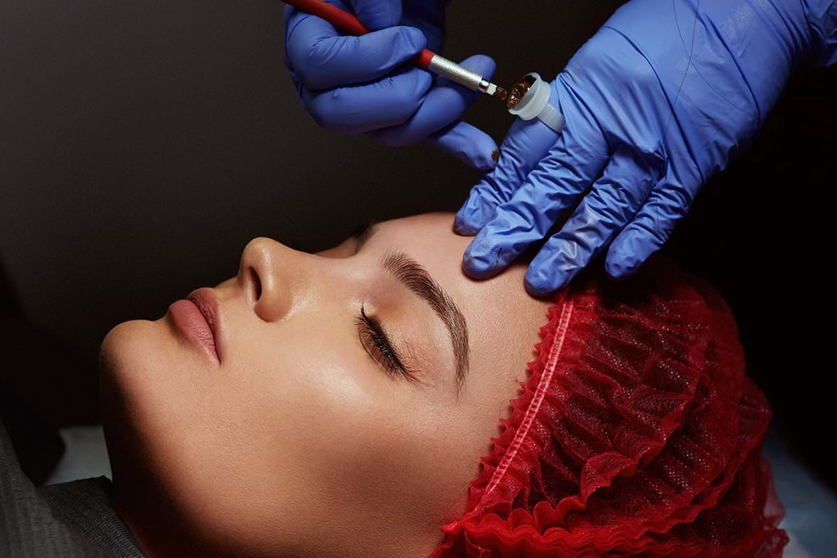 Person receiving eyebrow microblading; artist in blue gloves using a tool on the forehead.