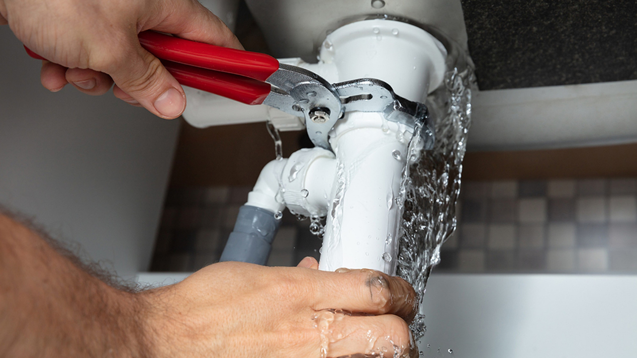 A person uses a wrench to tighten a pipe under a sink; water is leaking.
