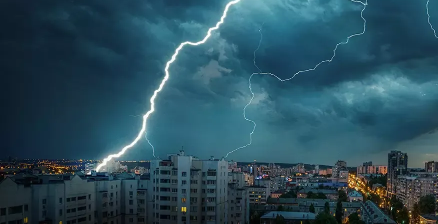 Lightning strikes a city skyline at dusk. Dark blue clouds, buildings, and bright white bolt.
