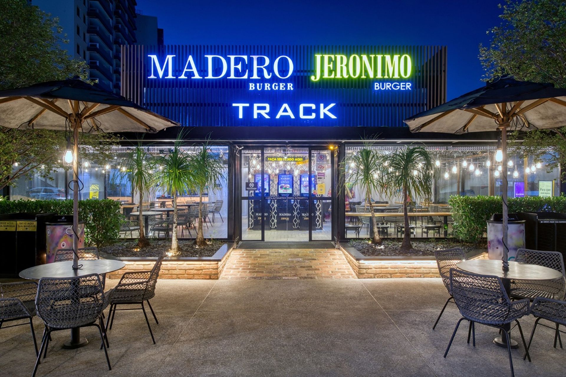 Madero Jeronimo Burger restaurant exterior at night. Neon sign, outdoor seating, glass front with visible patrons.