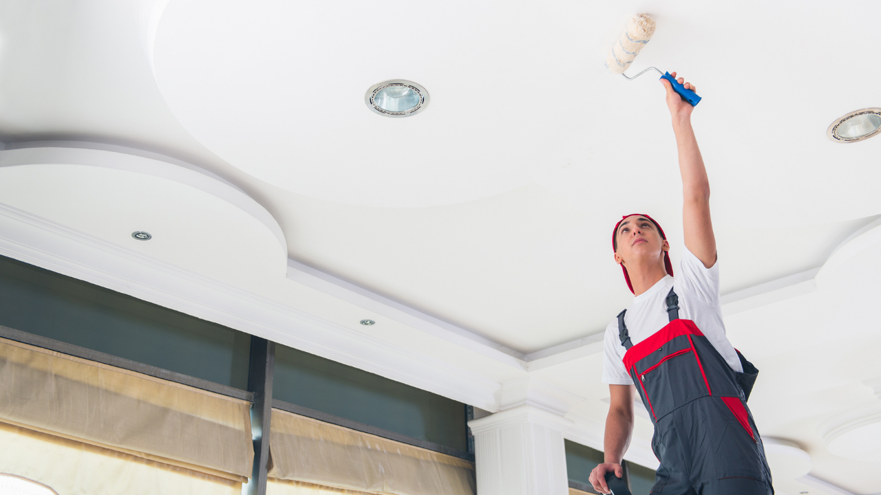 Person painting a white ceiling with a paint roller in a room with recessed lighting.