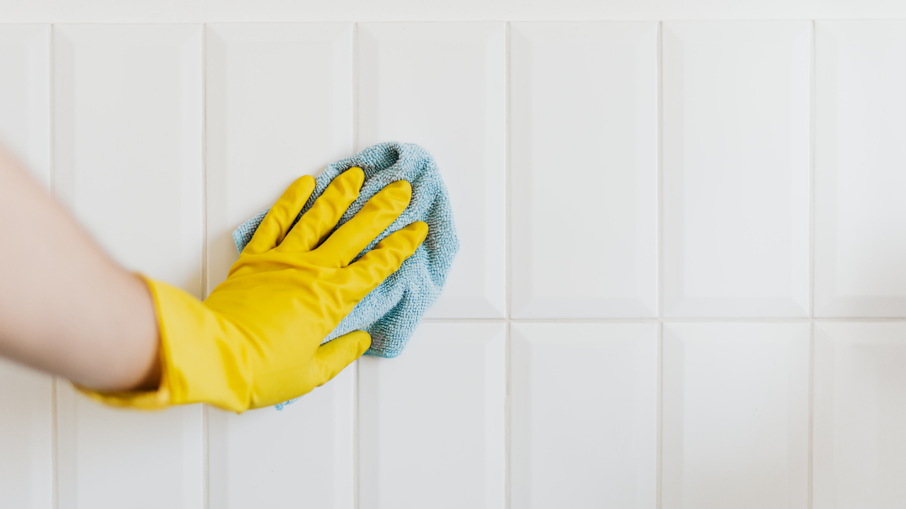 Hand in yellow glove wipes white tiled wall with blue cloth.