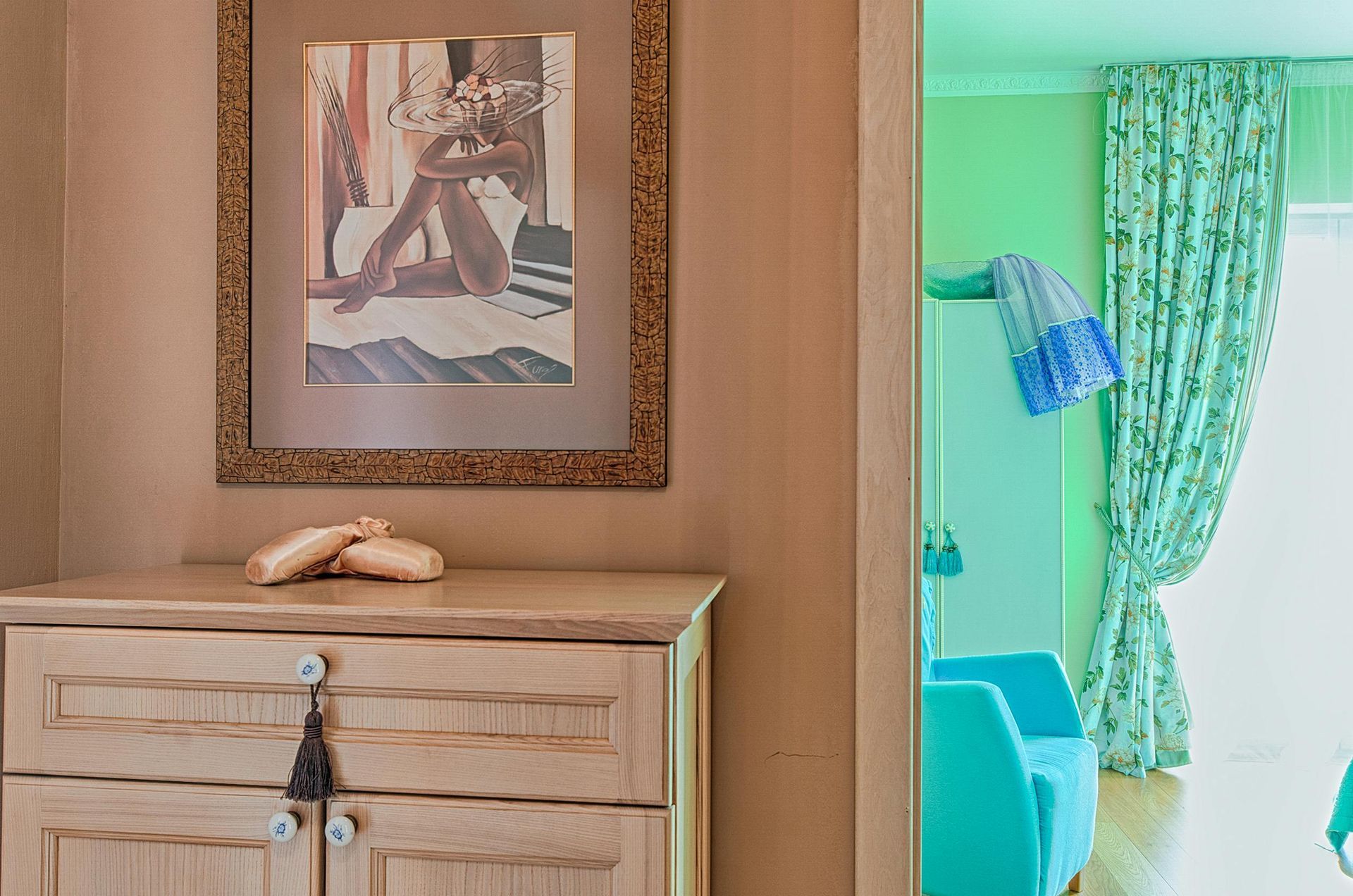 Wooden cabinet with framed art and ballet shoes. Open doorway reveals turquoise chairs and patterned curtains.