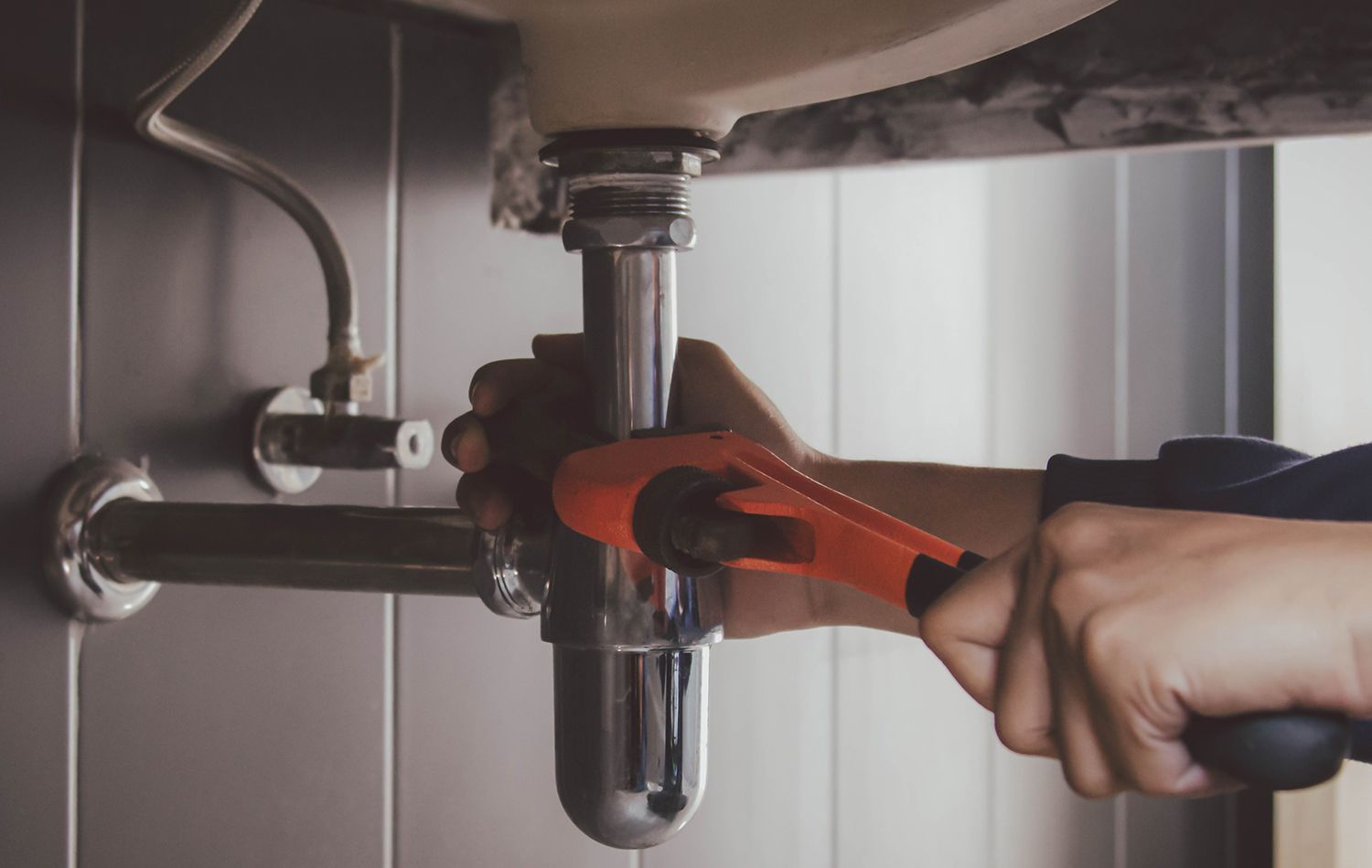 Hand using an orange pipe wrench to tighten a chrome sink drain pipe under a sink