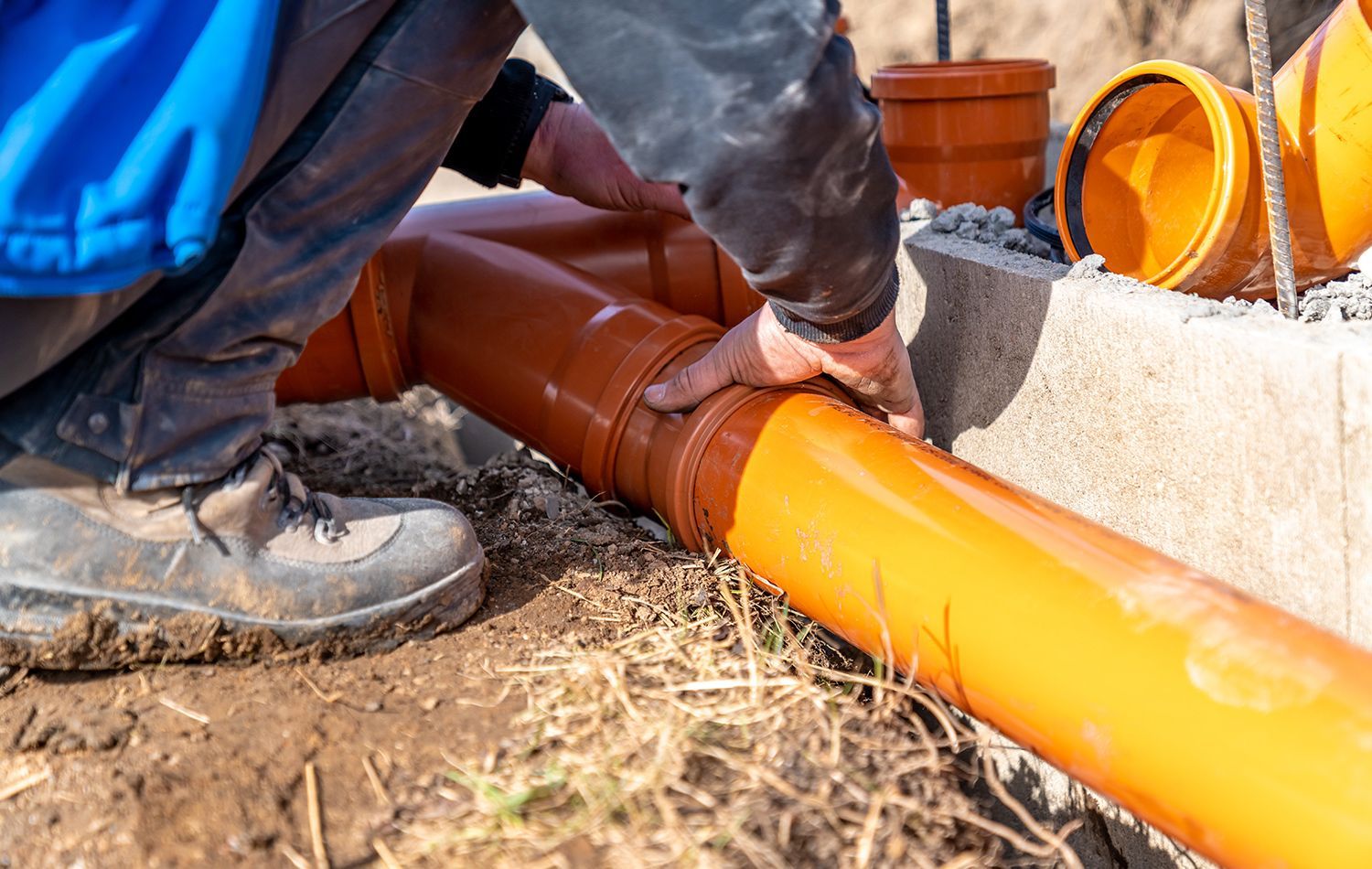 A plumber in work clothes kneeling in a bathroom, using a wrench to tighten the pipe underneath a wall-mounted sink.