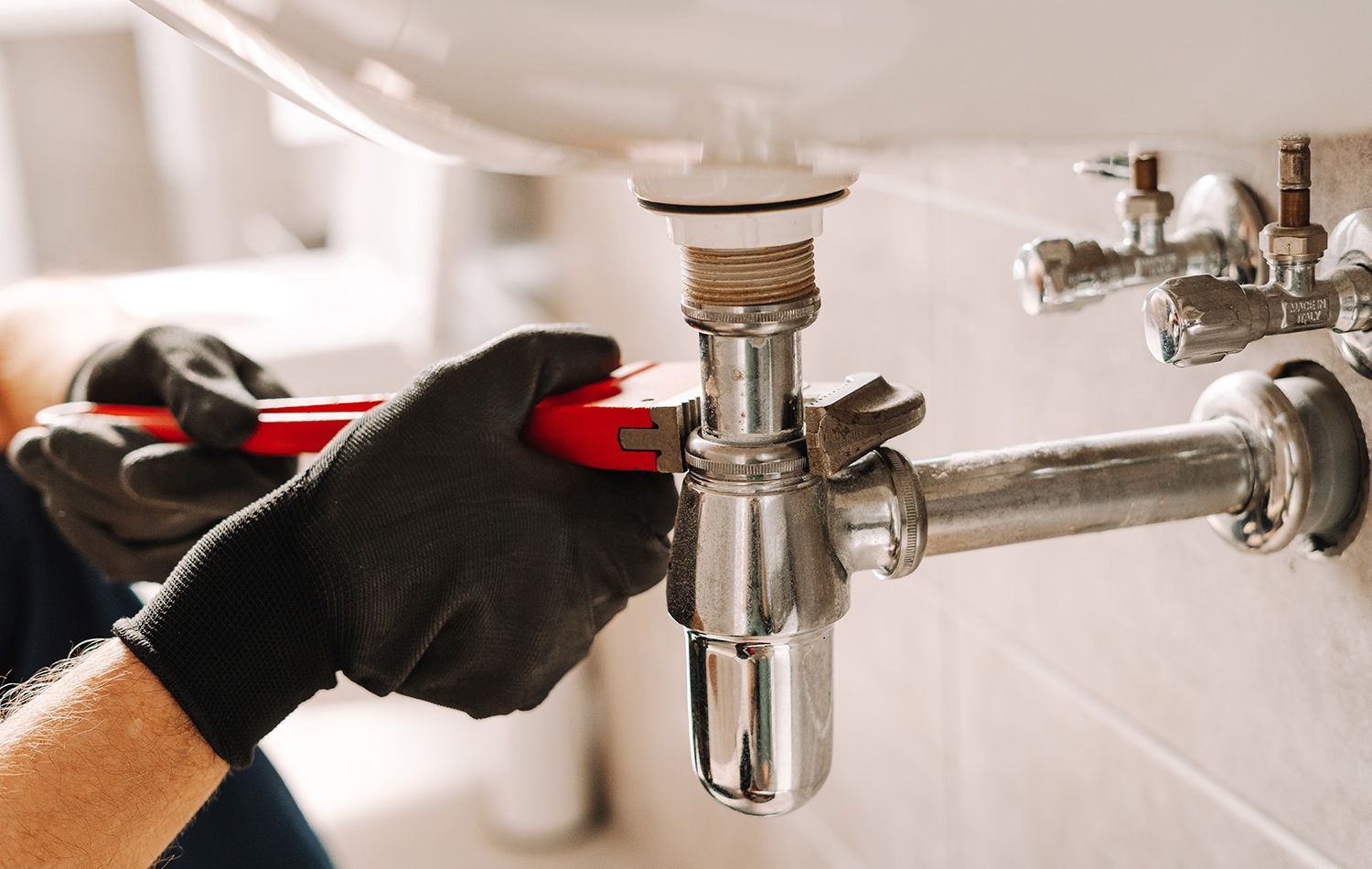 A plumber in work clothes kneeling in a bathroom, using a wrench to tighten the pipe underneath a wall-mounted sink.