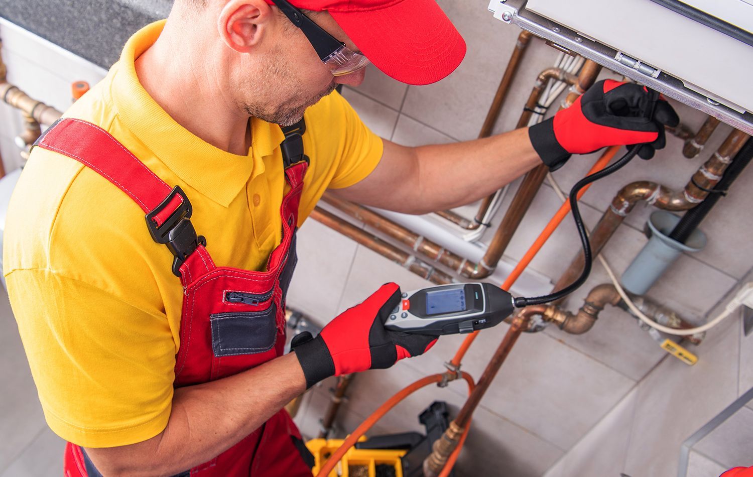 A plumber in work clothes kneeling in a bathroom, using a wrench to tighten the pipe underneath a wall-mounted sink.