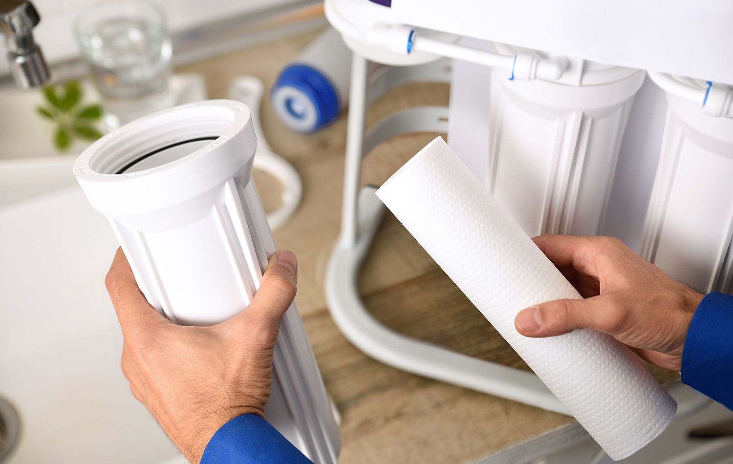 A plumber in work clothes kneeling in a bathroom, using a wrench to tighten the pipe underneath a wall-mounted sink.