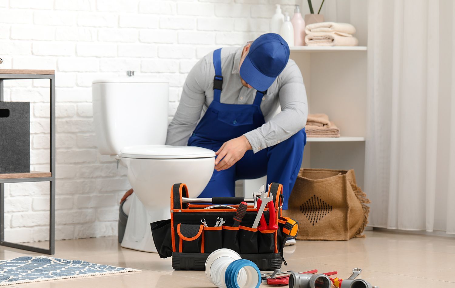 A plumber in work clothes kneeling in a bathroom, using a wrench to tighten the pipe underneath a wall-mounted sink.