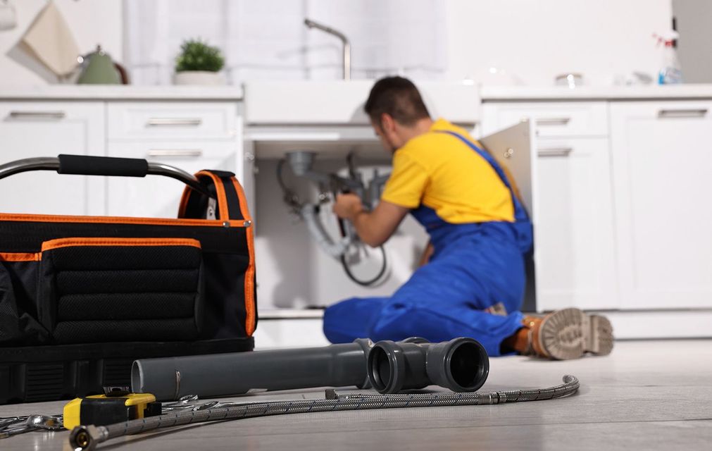 A worker wearing gloves installs or repairs a chrome P-trap pipe under a sink.