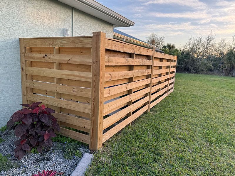 Wooden fence enclosing a corner of a house, protecting an AC unit, set on grass with a plant.