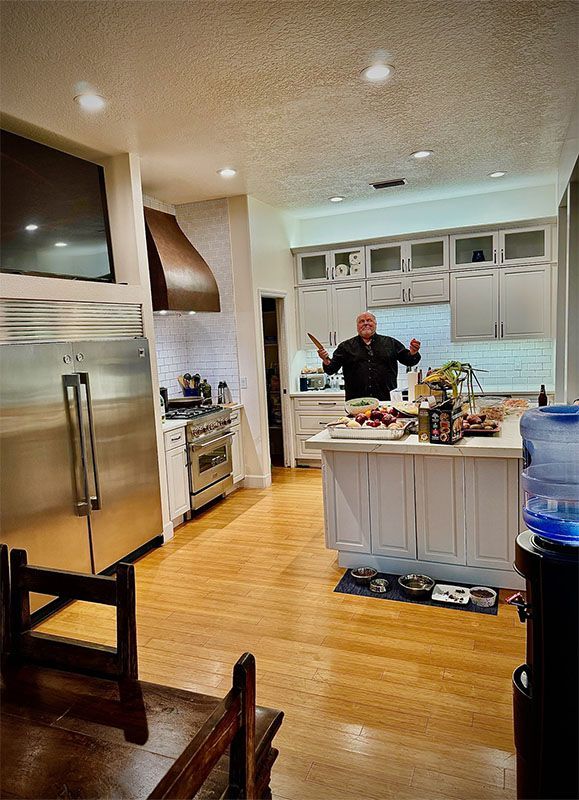 Man in kitchen with open arms, surrounded by food on island. Wood floors, white cabinets.