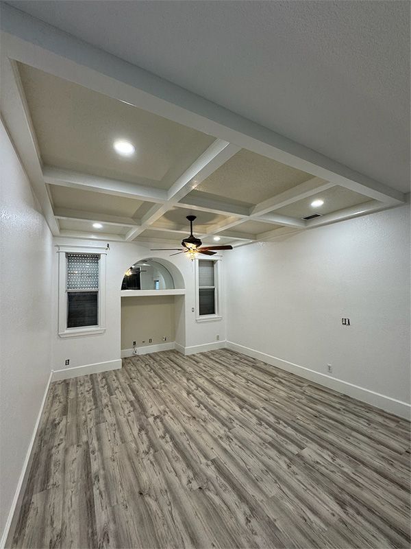 Empty room with wood-look floors, white walls, and a coffered ceiling. Windows and a recessed archway are visible.