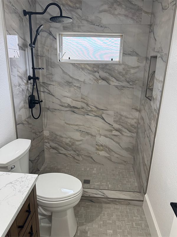 Bathroom with marble-look tiled shower, black shower fixtures, toilet, window, and small tile floor.