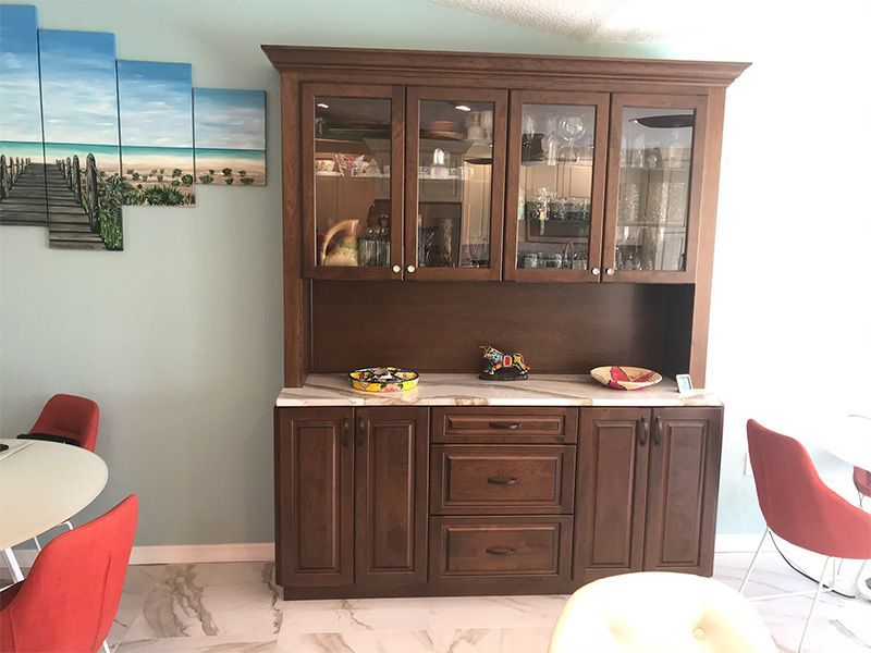 Wooden hutch with glass doors, marble countertop, and lower cabinets. Painted beach art on the wall and red chairs.