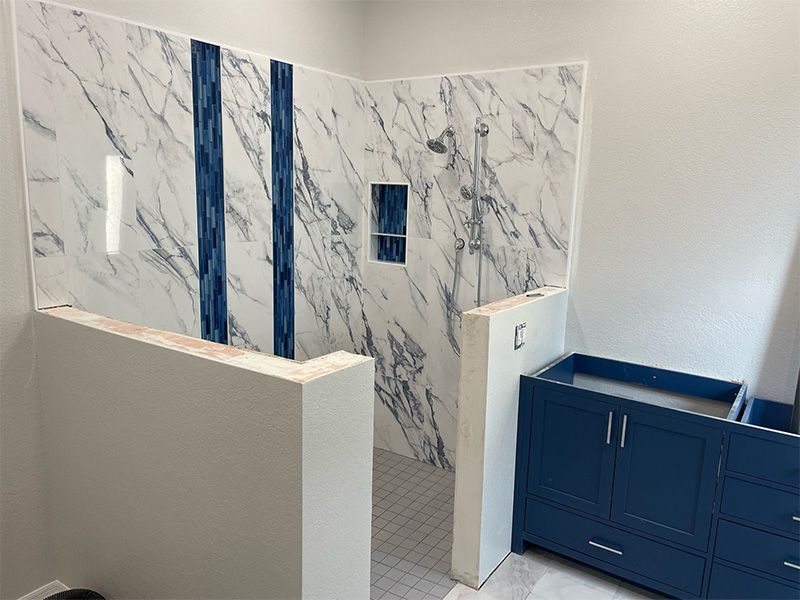 Bathroom under construction with marble-look shower walls, blue accent strips, a blue vanity, and gray walls.