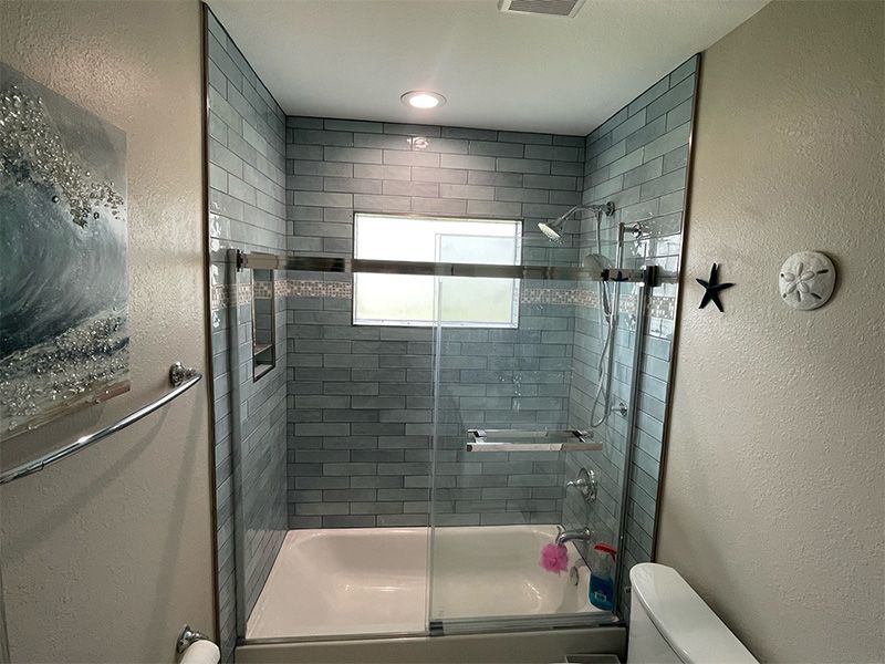 Bathroom with blue tiled shower, white tub, and glass door. Includes decor like artwork and starfish.