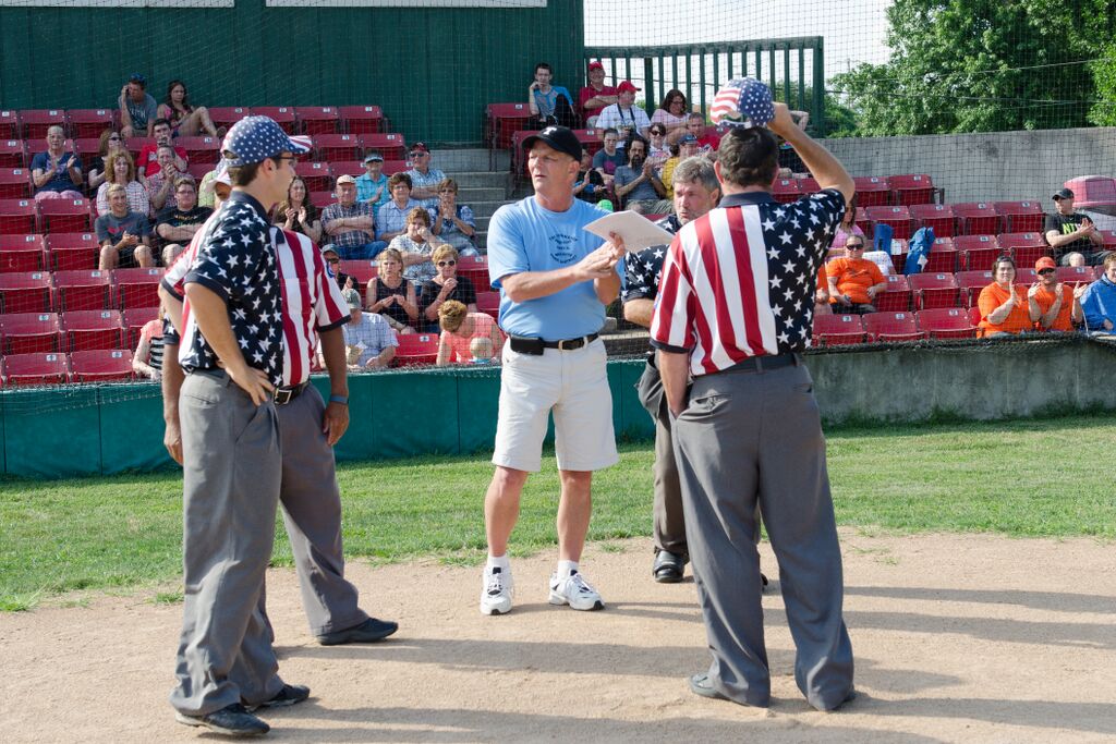 Baseball umpires, American flag shirts, talking with a man in white shorts on a field, bleachers in background.