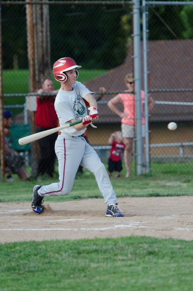 Baseball player swinging bat, hitting a ball; grassy field, spectators in background.