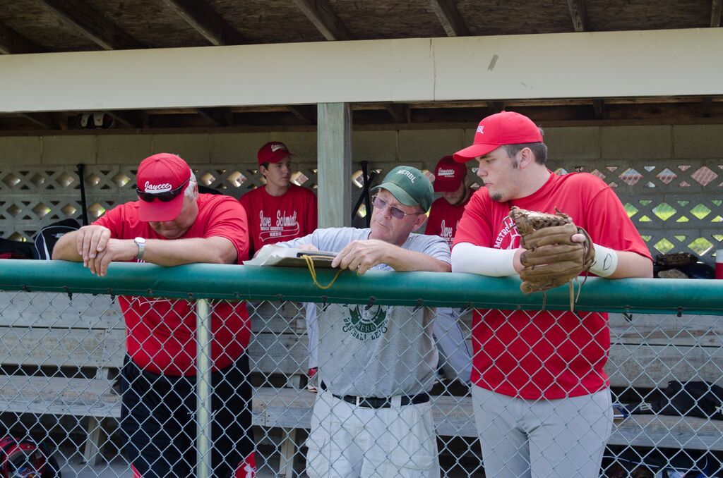 Baseball players and coach in dugout, discussing plays. Red shirts, hats, and glove. Green railing.