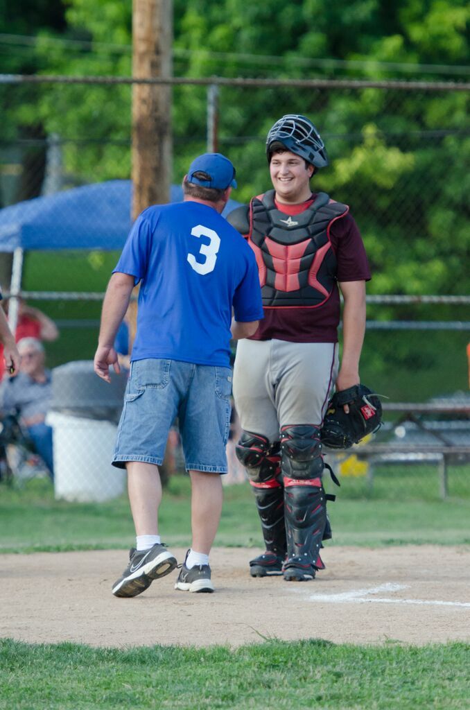 A baseball coach in blue shirt shakes hands with catcher in full gear. They are on a baseball field.