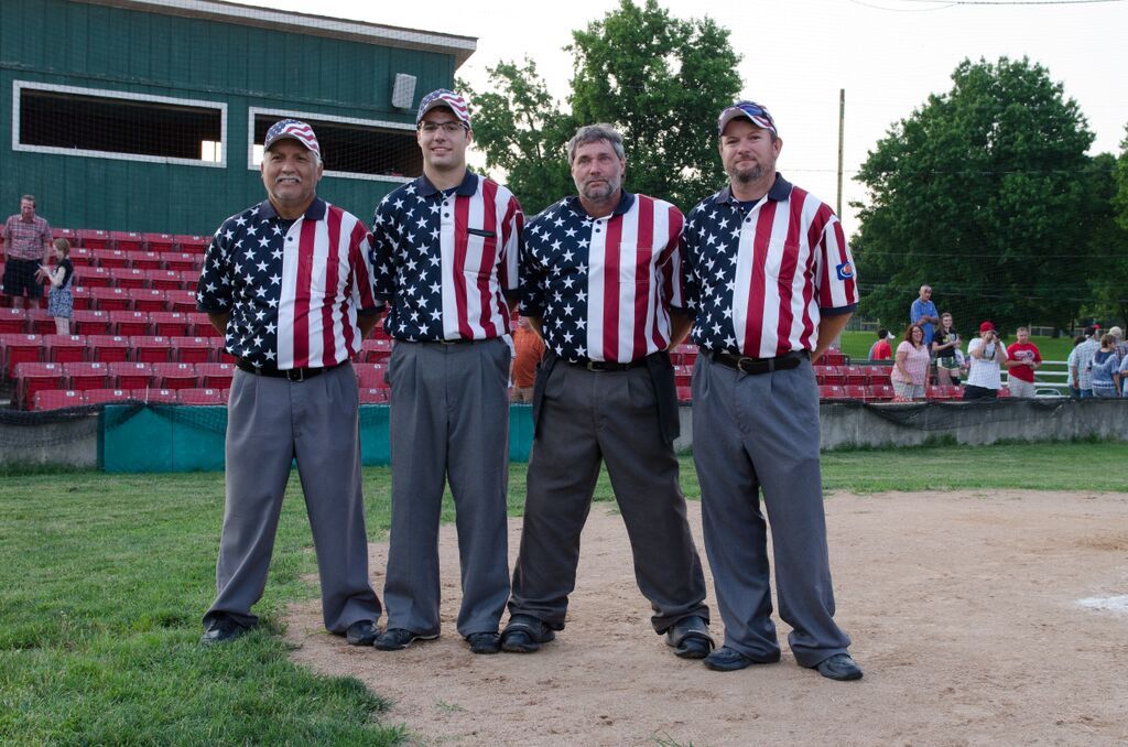 Four baseball umpires in American flag-themed shirts stand on a baseball field.