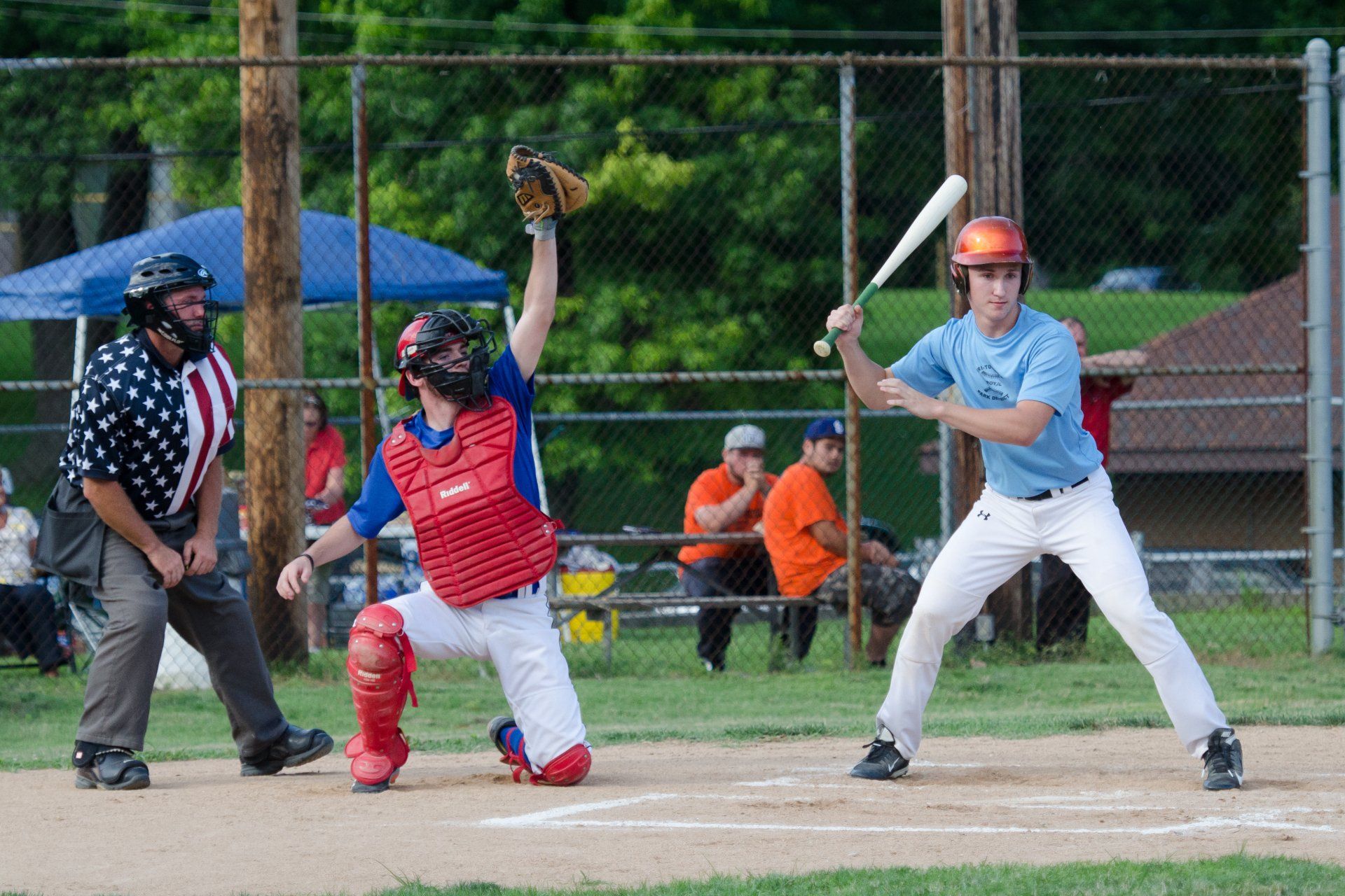 Baseball game: Batter at plate, catcher in crouch, umpire with American flag shirt, spectators in background.