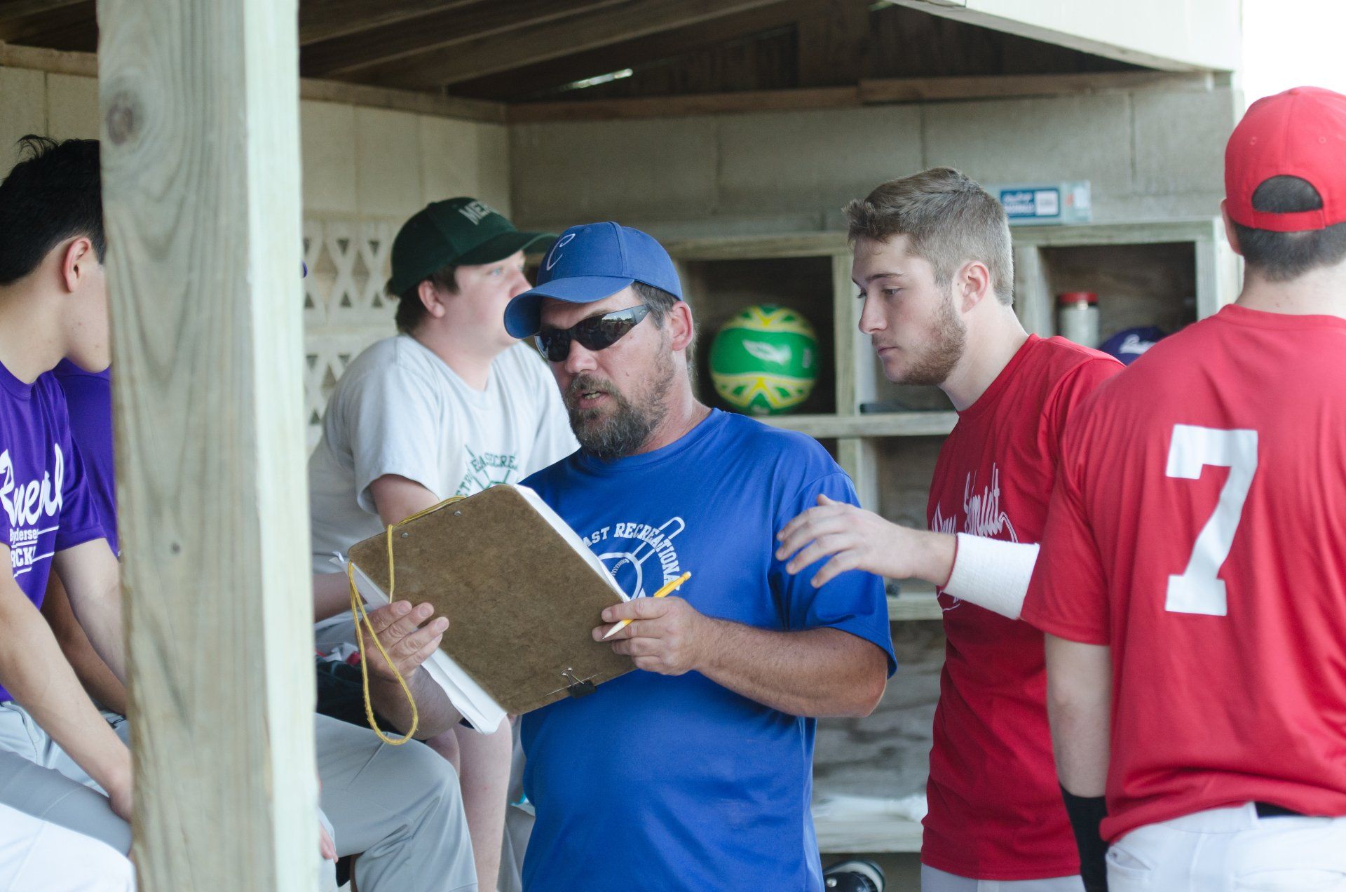 Baseball coach in blue discusses strategy with players in dugout; they wear red and purple uniforms.