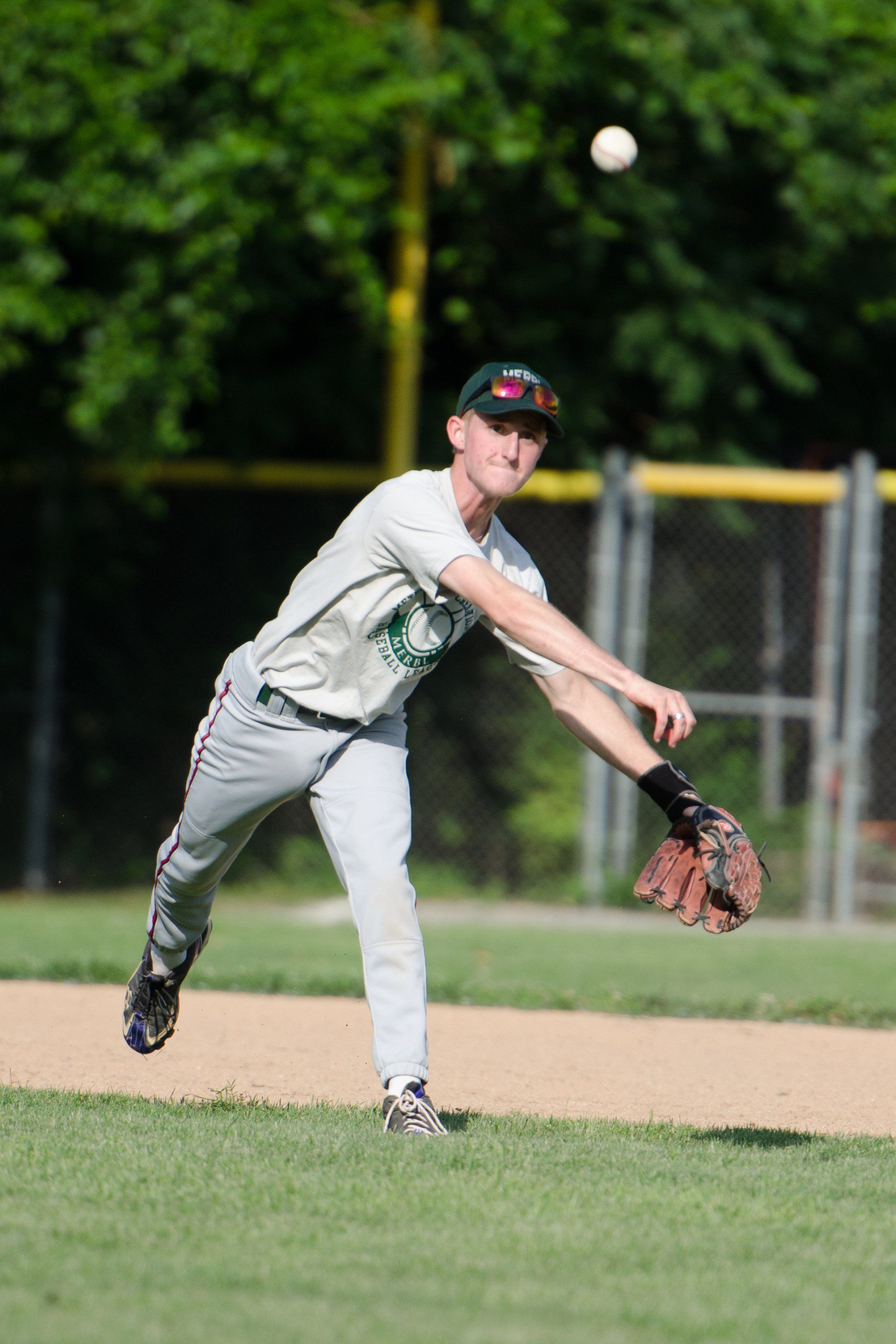 Baseball player throwing ball on field. Wearing gray pants, green shirt and hat, with glove.