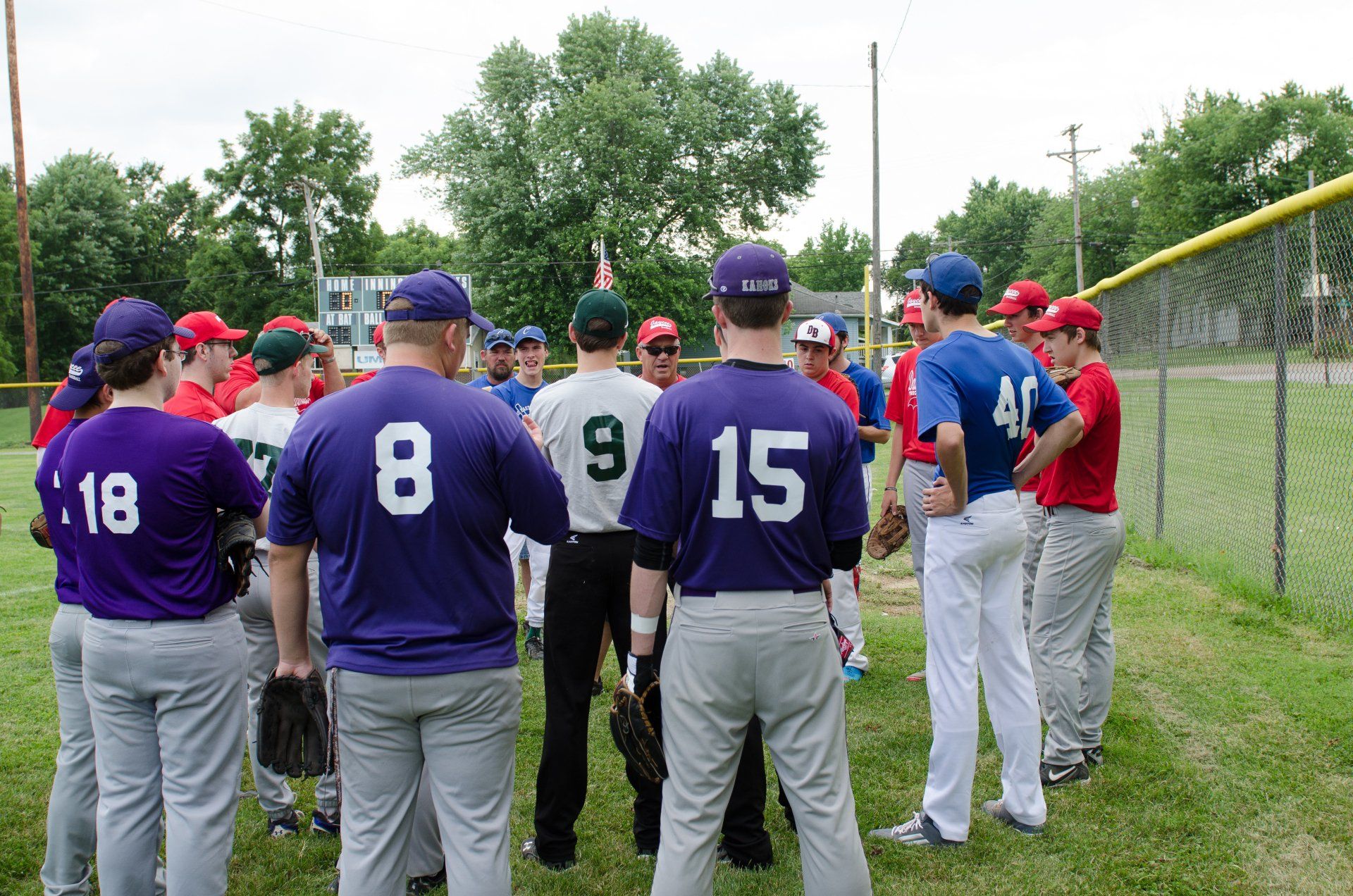 Baseball teams in huddle on a field. Players in purple and red jerseys gather around an umpire.